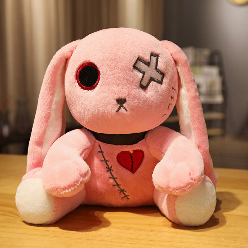Pink Dark Bunny plushie sitting upright on a wooden table, front-facing close-up shot showing stitched heart detail, crossed eye embroidery, and floppy bunny ears, soft gothic plush toy with pastel pink fabric and embroidered accents, cute alternative plush decor.