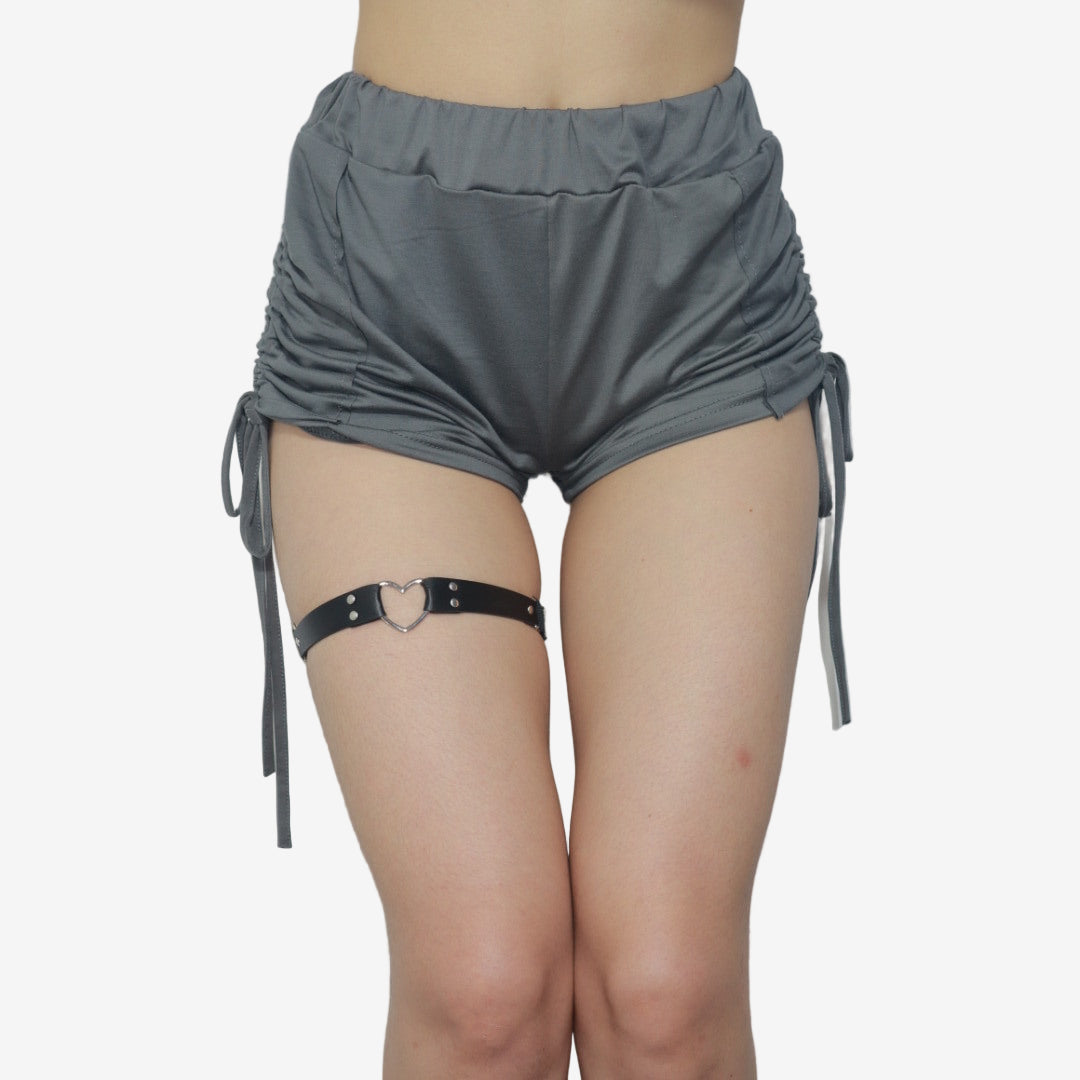 Full-body view of a femboy wearing a black heart-shaped leather garter paired with loose gray shorts. The minimalist design complements the femboy fashion aesthetic.