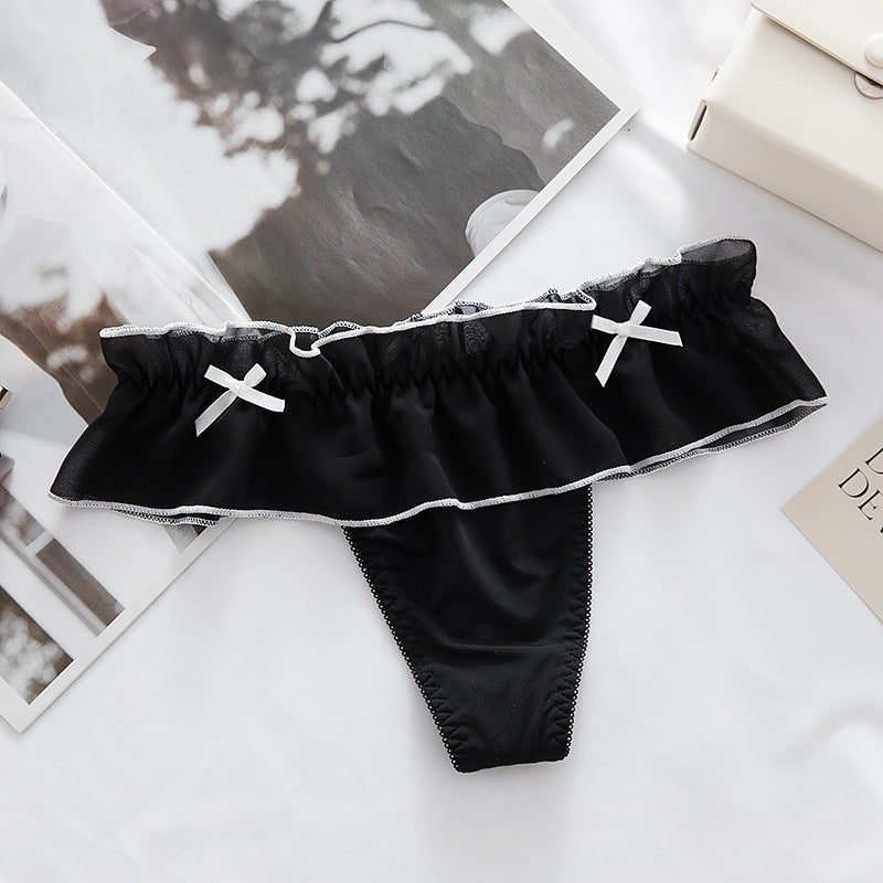 Overhead flatlay photo of black ruffled femboy maid panties with white accent bows, styled on a minimal white setup for a cute femboy underwear look.