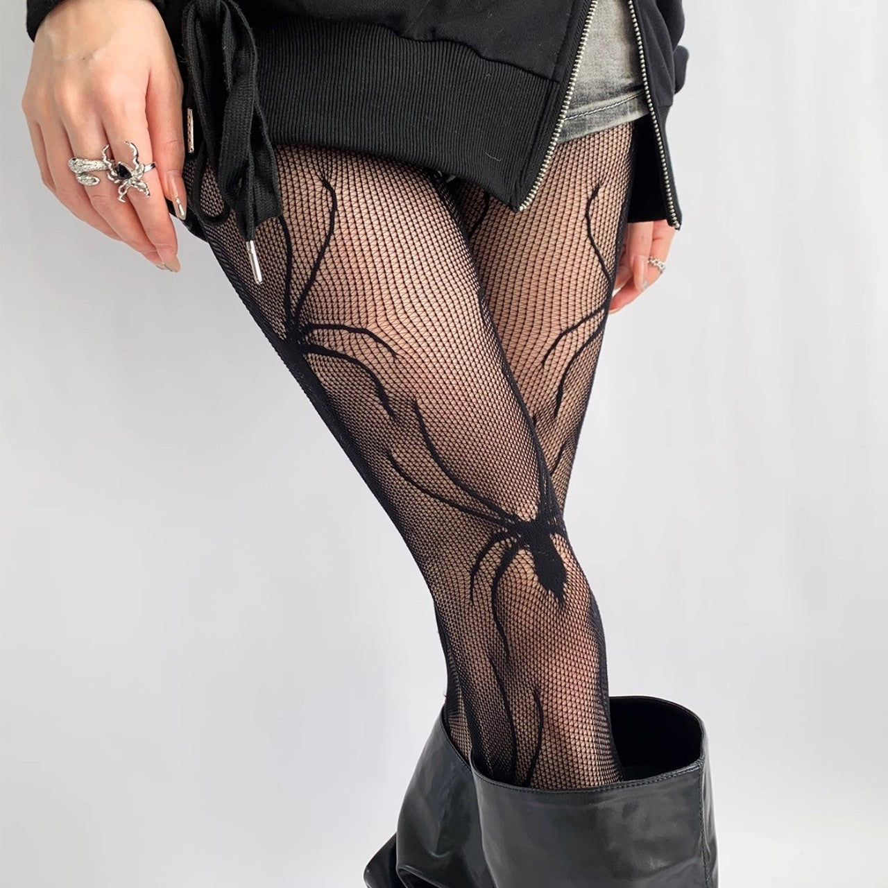 three-quarter close-up shot of a model’s legs in black spider-pattern mesh pantyhose paired with black ankle boots, emphasizing the intricate spider motif against a light gray floor for femboy outfit ideas.