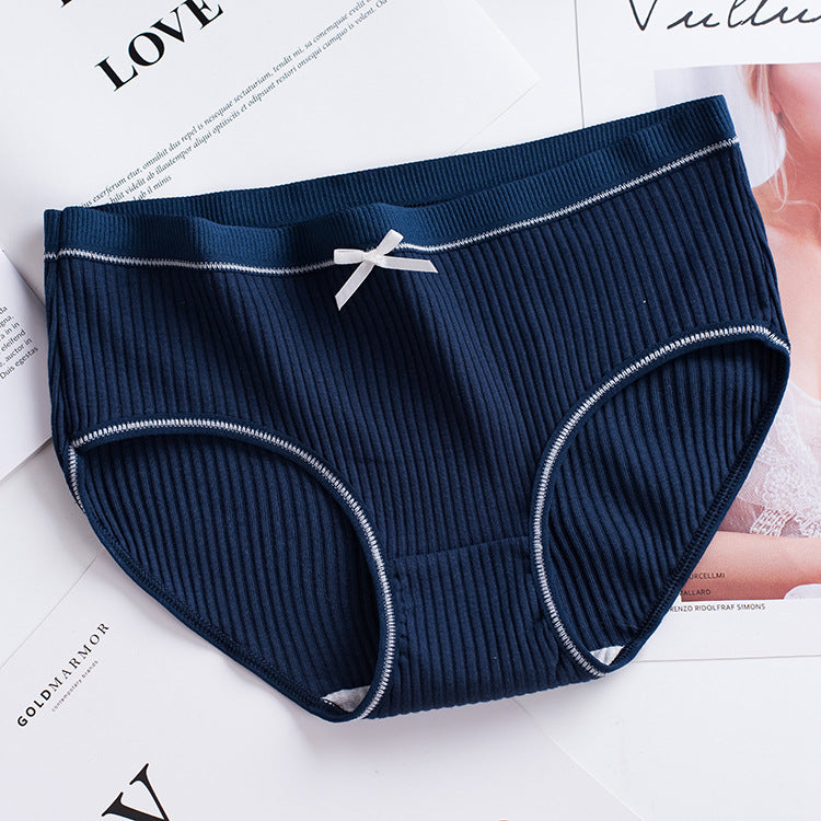 overhead shot of a navy ribbed brief with white contrast stitching and a small white bow, laid on a white tabletop with magazine pages, showcasing femboy underwear essentials.