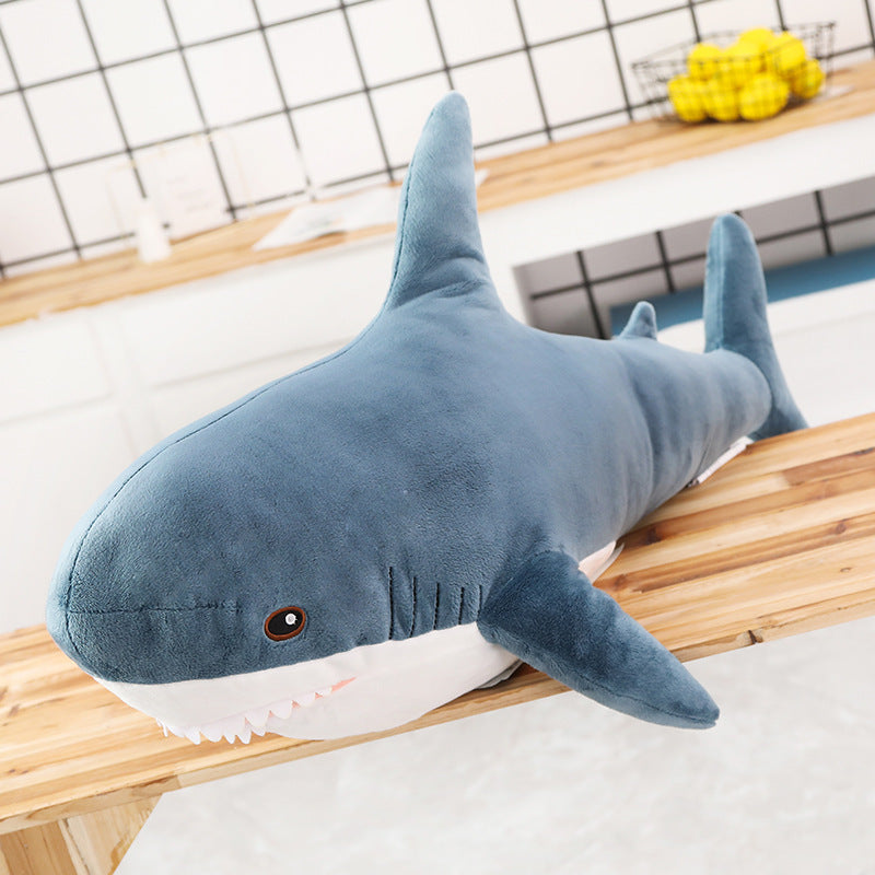 Close-up angled view of large blue Femboy Blahaj pillow plushie, oversized cuddly shark plush toy displayed in cozy home interior, wooden tabletop background