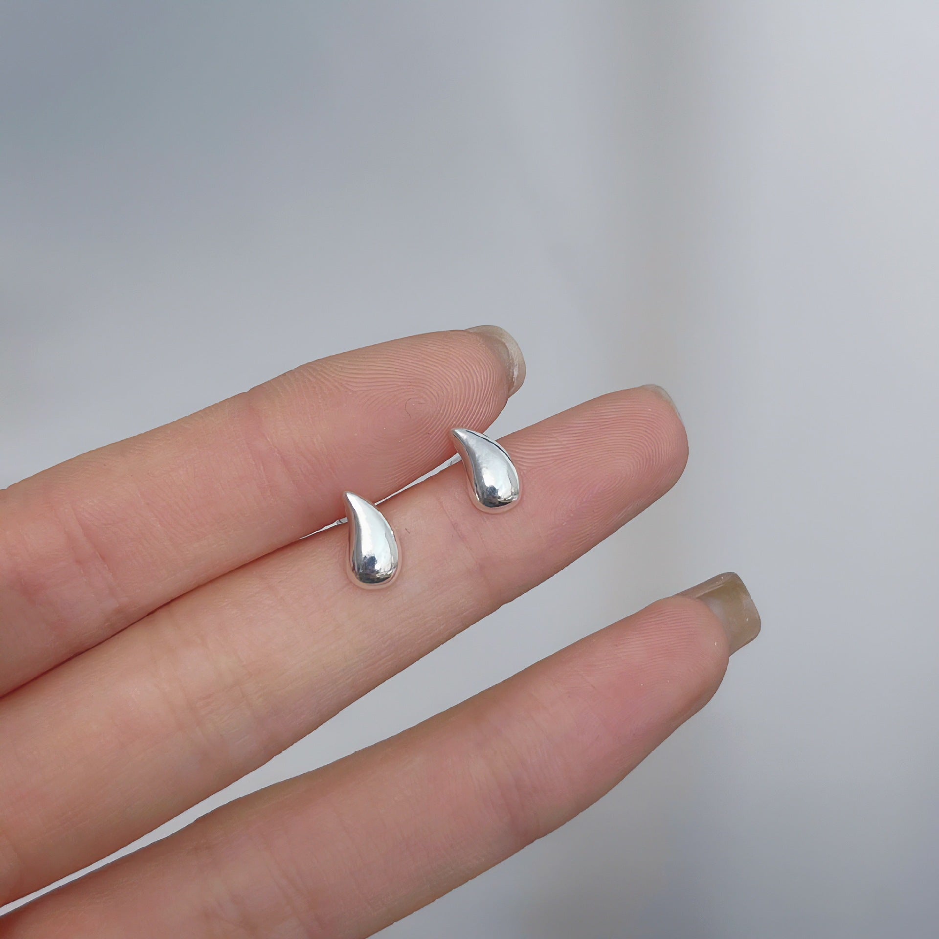 Close-up of silver teardrop-shaped earrings on a hand, showcasing minimalist and femboy fashion design.