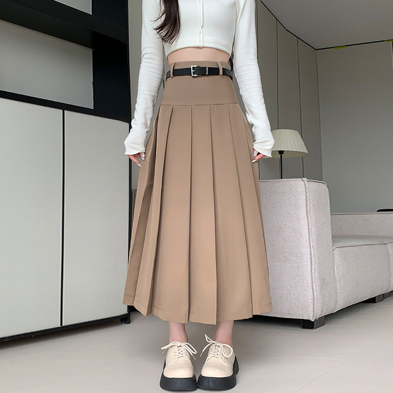 Medium shot of a model wearing a beige midi pleated skirt with a high waist and belt detail, embodying femboy fashion style, captured indoors with minimalistic decor in the background.