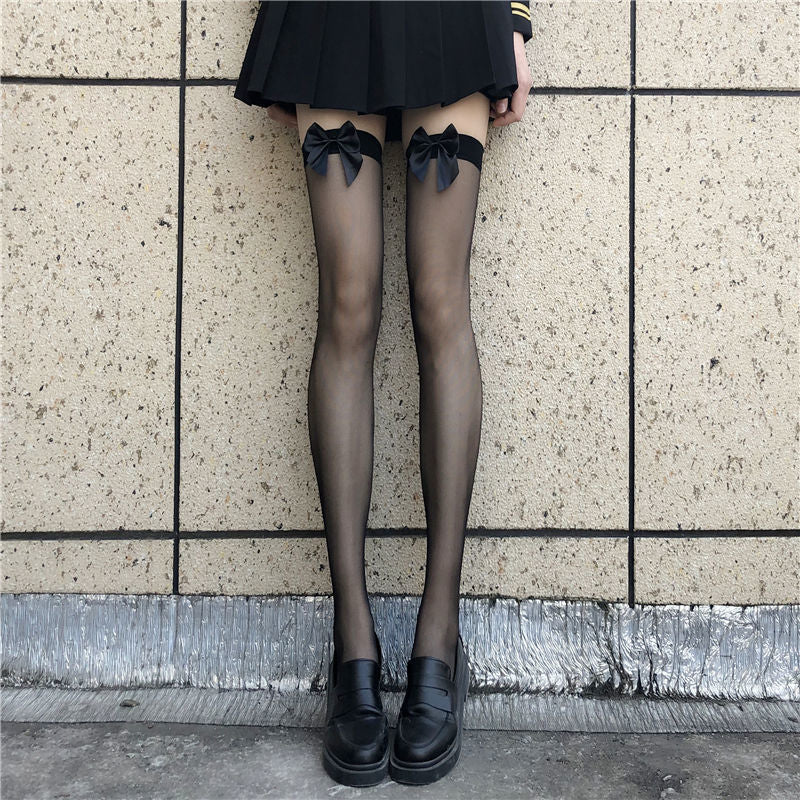 A front view of black sheer stockings with satin bows, worn with a black pleated skirt against a neutral outdoor wall. The stockings’ delicate bow detailing adds an elegant touch, perfect for enhancing femboy outfits with playful, feminine femboy clothing.