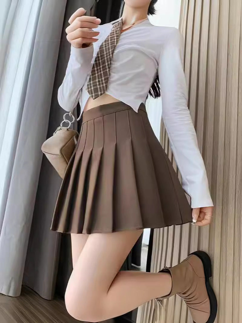 A close-up of a model dressed in a brown pleated skirt with a white top and plaid tie, set against a neutral indoor background. The pleats in the skirt add volume and structure, ideal for styling sophisticated femboy outfits for a refined femboy fashion sense.