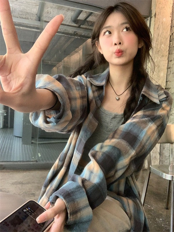 A playful selfie of a person in a blue plaid button-up shirt, making a peace sign, seated in a cozy café. The image portrays the laid-back vibe of femboy fashion.