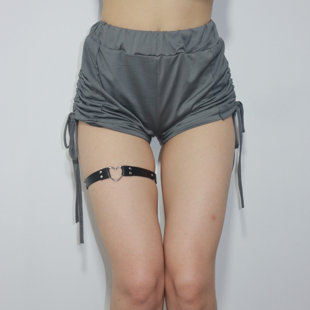 Full-body view of a femboy wearing a black heart-shaped leather garter paired with loose gray shorts. The minimalist design complements the femboy fashion aesthetic.