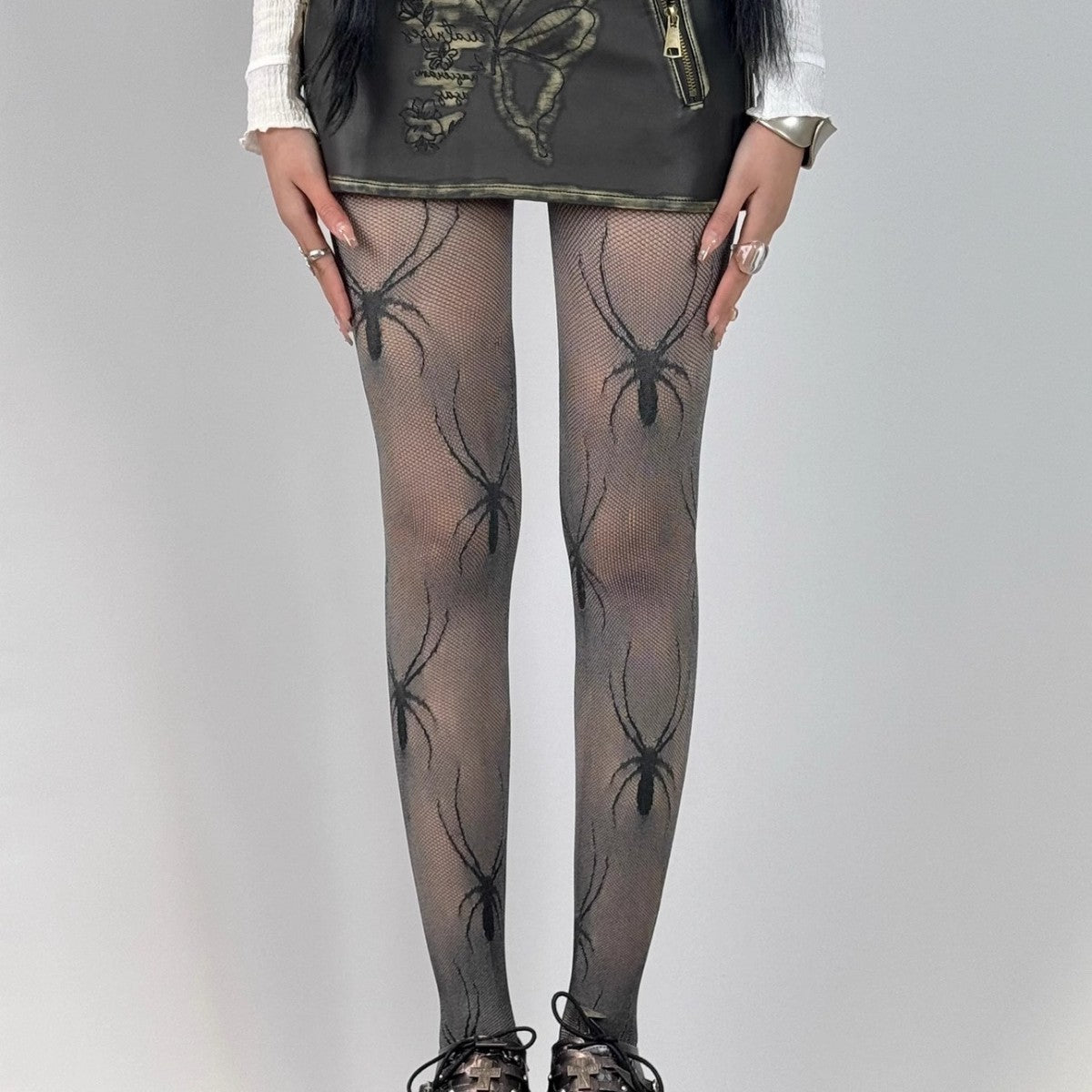 eye-level medium full-body shot of a model wearing black spider-print mesh pantyhose with chunky black platform heels and a leather mini skirt in a gray-walled room, perfect for alternative femboy styling.
