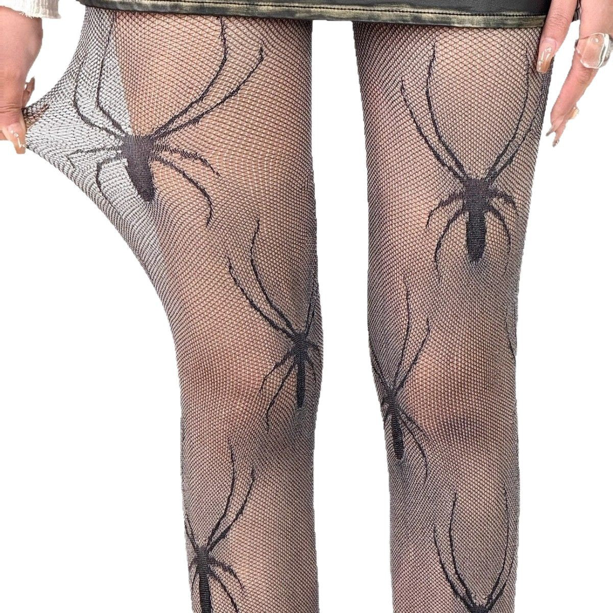 close-up flat-front shot of black spider-print mesh pantyhose being gently stretched by a model’s hand, shown under a distressed mini skirt against a white background, highlighting an edgy femboy look.

