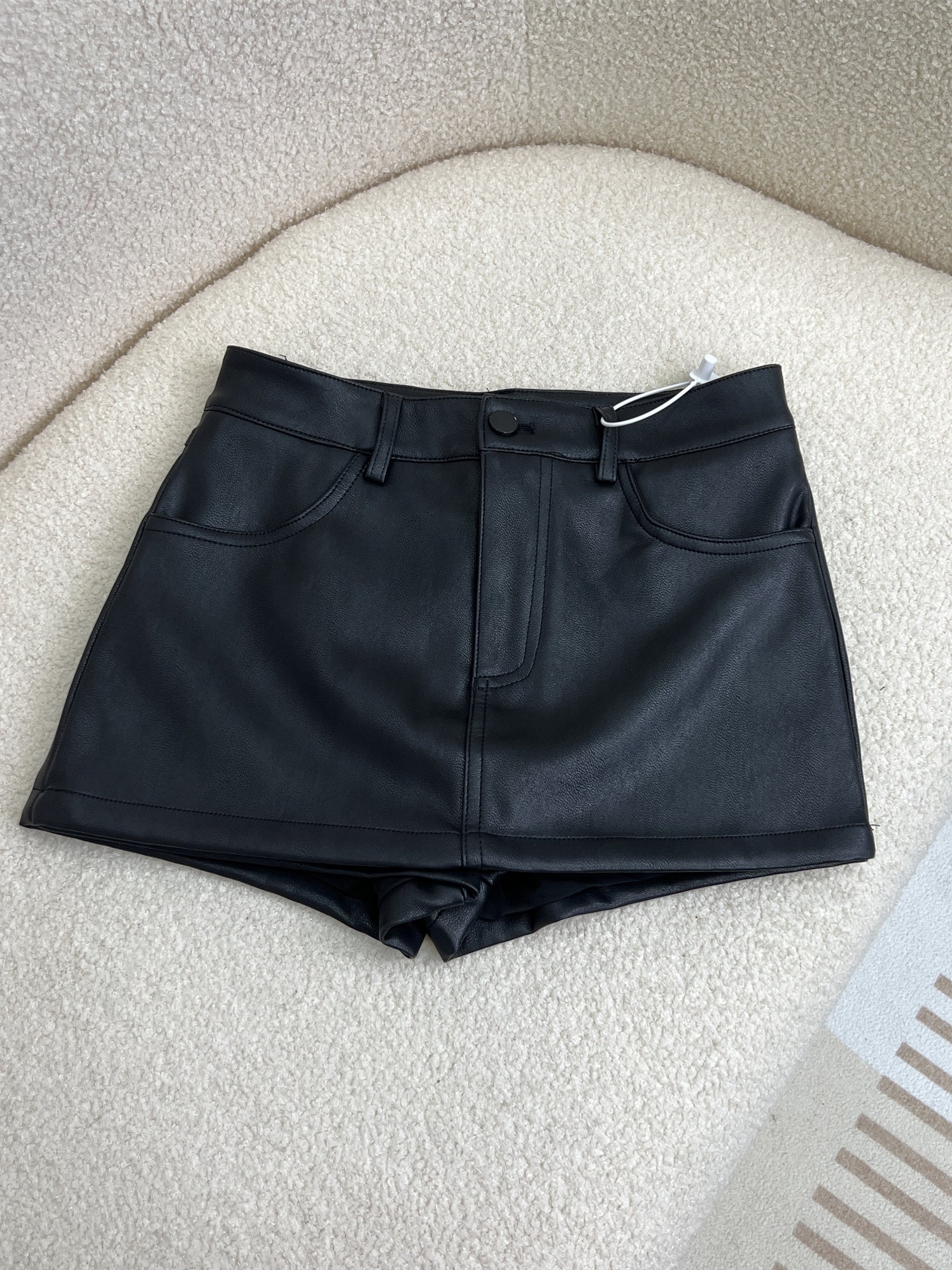 Flat lay product image of a black PU leather femboy skirt with built-in shorts, featuring a smooth faux leather finish, front button closure, belt loops, and front pockets, photographed on a soft cream fabric background, minimalist studio lighting, showcasing femboy clothing and femboy fashion.