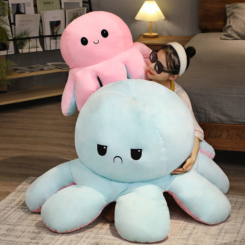 Wide lifestyle shot of oversized reversible octopus plushies in blue, pink, and orange displayed with a seated model, photographed from a slightly elevated front angle in a cozy living room setting, emphasizing scale, softness, and playful design from a femboy shop.