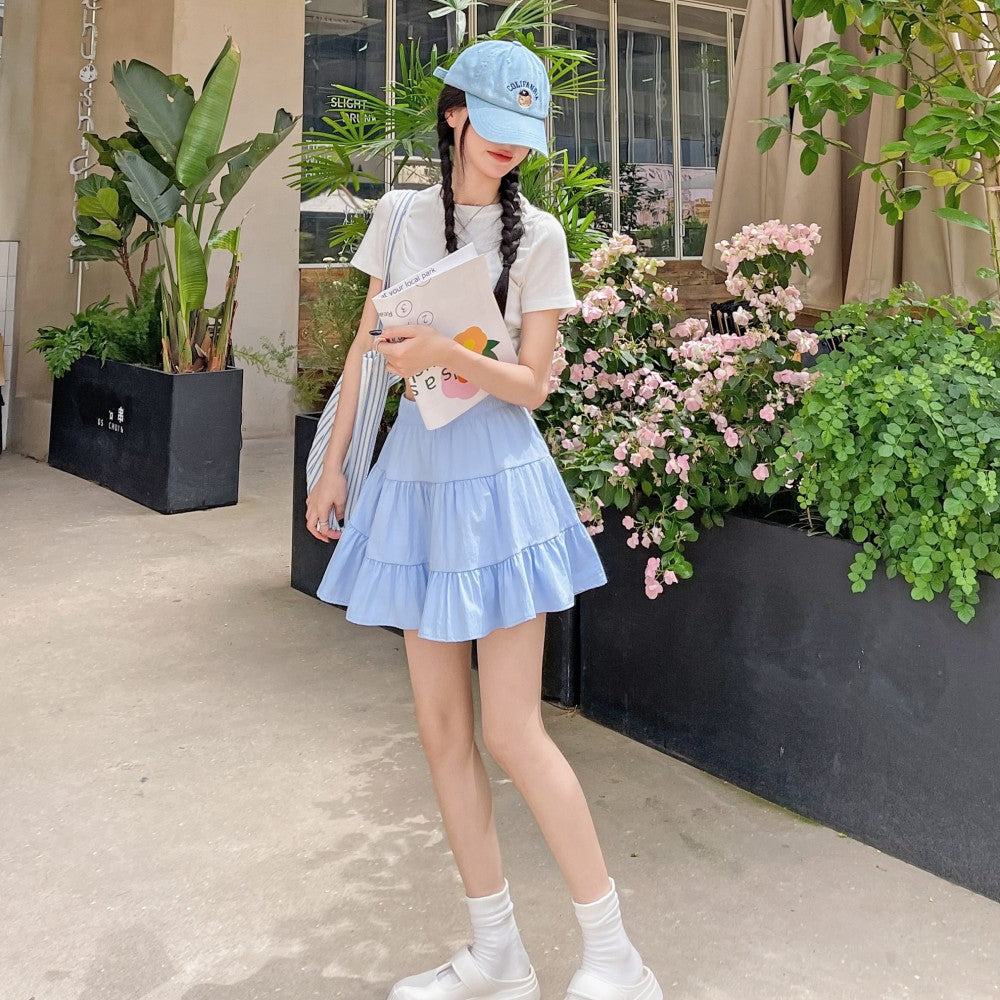 Femboy in light blue outfit standing outdoors with plants and flowers in the background