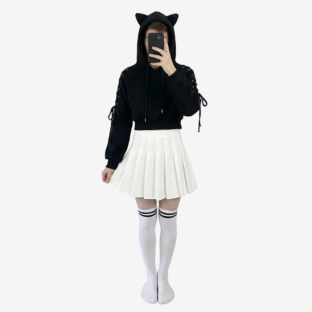 Person wearing a black hoodie with cat ears, white pleated skirt, and white socks with black stripes in front of a mirror.