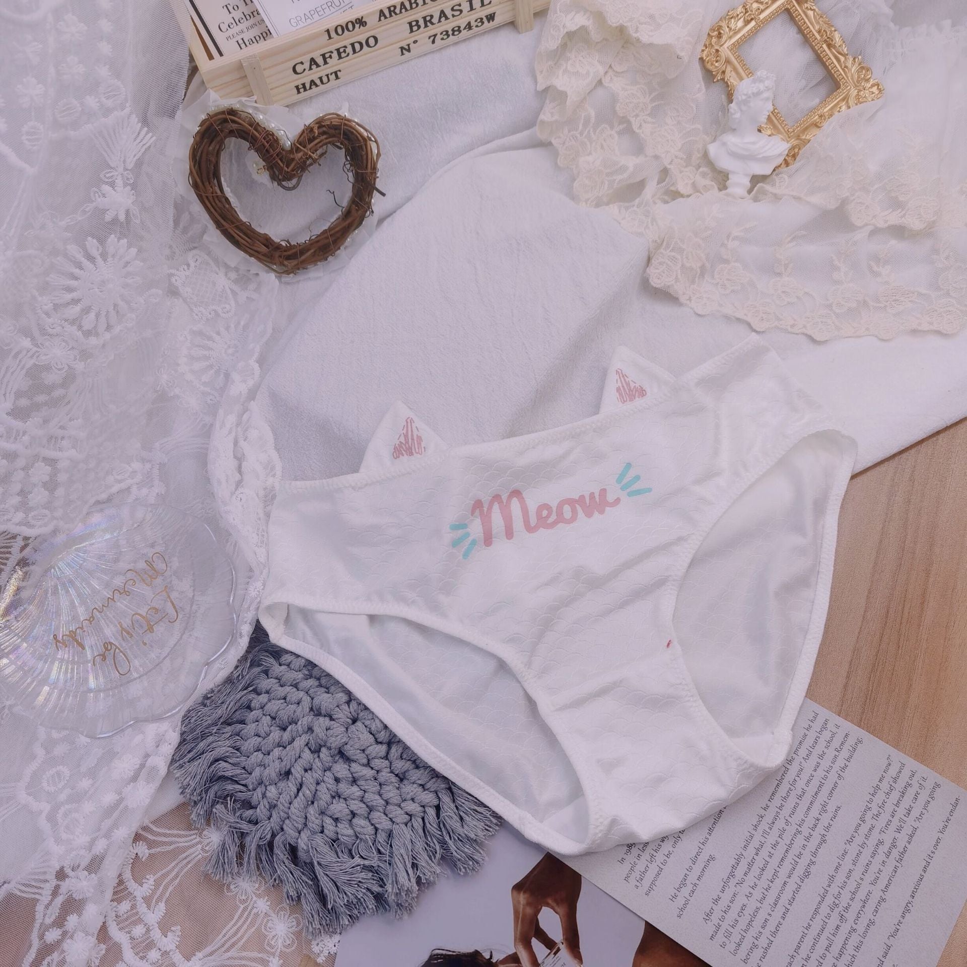 White cat pouch femboy panties with meow graphic and cat ear waistband detail, styled flat lay with lace fabric background