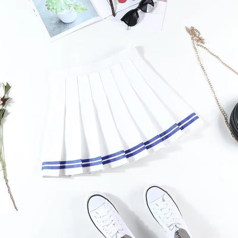 Top-down close-up shot of a white pleated skirt with blue stripes, laid on a white surface alongside accessories. The minimalistic background enhances the bright, fresh look of this femboy clothes staple, perfect for femboy fashion and casual femboy outfits.